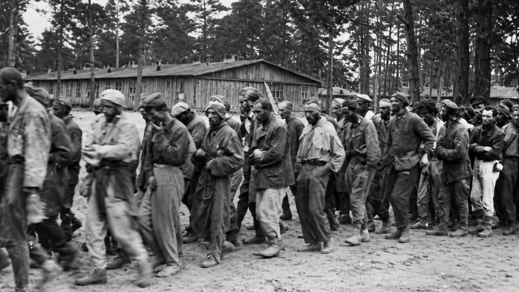 Історична чорно-біла фотографія часів Другої світової війни. Група виснажених радянських військовополонених у літній та зимовій формі йде колоною по території нацистського концтабору у Дарниці. На задньому плані видно дерев'яні табірні бараки та сосновий ліс.