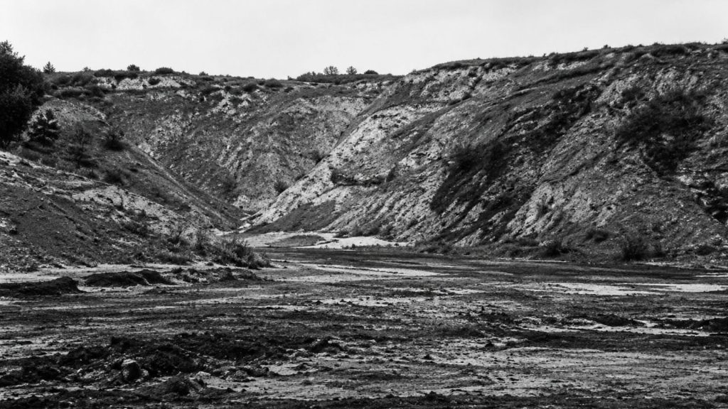 Історичне чорно-біле фото. На передньому плані видно широке дно Бабиного Яру з темною, випаленою та розритою землею, вкритою попелом і залишками вогнищ. На задньому плані височіють круті, обривисті схили яру зі світлої глини та піску, що утворюють природну стіну урочища під похмурим небом.