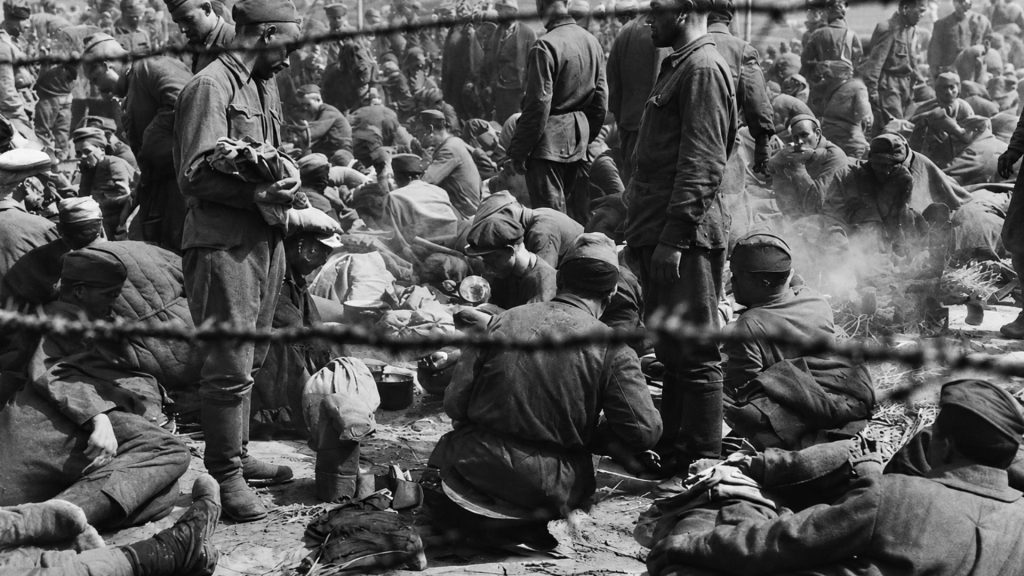 Історичне чорно-біле фото. Велика група виснажених радянських військовополонених у табірному секторі, знята крізь ряди натягнутого колючого дроту на передньому плані. Частина військових сидить на землі, інші стоять у затісному просторі. Подекуди з казанків або вогнищ здіймається дим. На задньому плані видніється велике скупчення людей та силуети військовополонених.