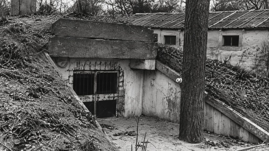 Історичне чорно-біле фото. По центру знято вхід до заглибленого в землю бетонного бункера (доту), який нацисти використовували як табірний карцер. Вхід закритий масивними залізобетонними балками, а дверний отвір перекритий міцними залізяками у вигляді решітки. На передньому плані праворуч стоїть стовбур дерева, а на задньому плані видніється дерев'яний одноповерховий табірний барак із вікнами.