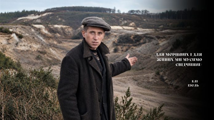 Чорно-білий скріншот із документальної кінохроніки з вертикальними лініями перешкод плівки. По центру великим планом зафіксовано Юхима Вілкіса — уцілілого свідка нацистських злочинів. Він одягнений у темне довге пальто та кепку, його обличчя напружене, а ліва рука піднята у жесті, що вказує назад на панораму глибокого й розлогого Бабиного Яру. На передньому плані праворуч внизу видно металеву деталь кінематографічного або військового обладнання.